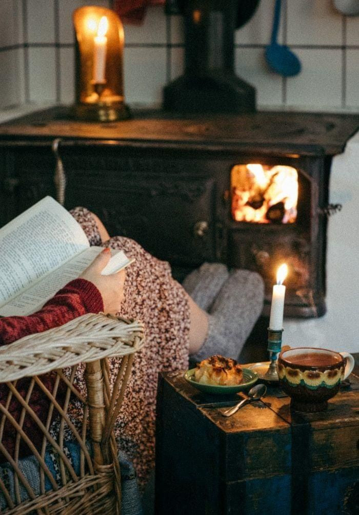 Cozy indoor scene with a person reading by a wood-burning stove, surrounded by candlelight, a hot drink, and a pastry on a rustic wooden table.