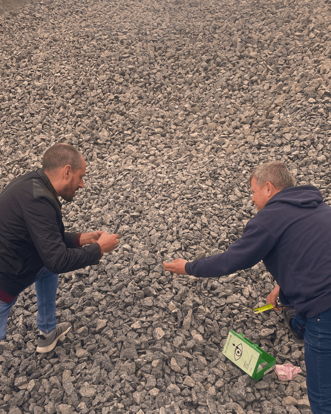 Two men crouching on a rocky surface, appearing to examine or collect small stones. One holds a green box with a warning label, and the other has a yellow tool or object in hand.
