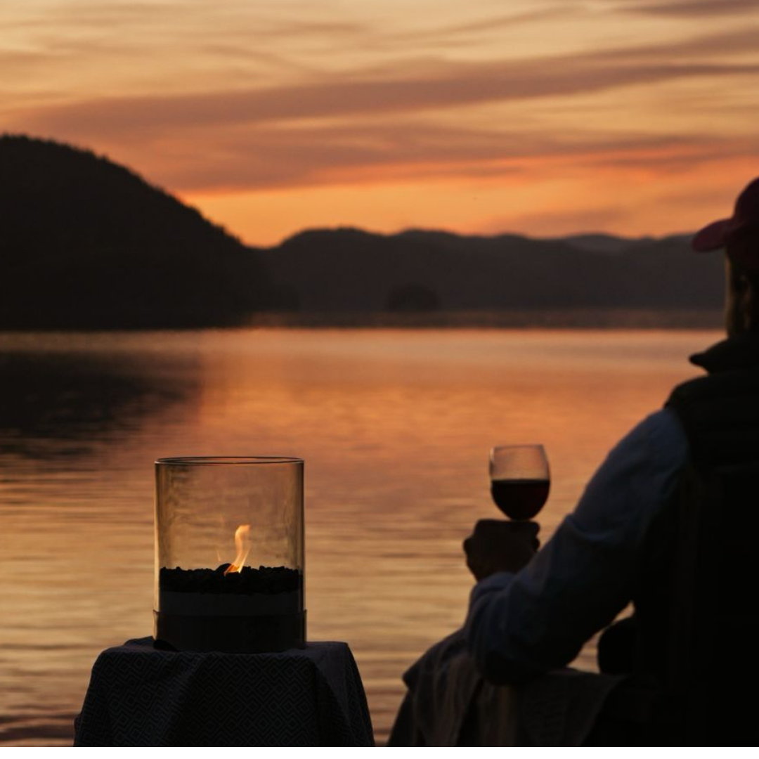 Man enjoys summer sunset by lake with real flame and glass of wine.