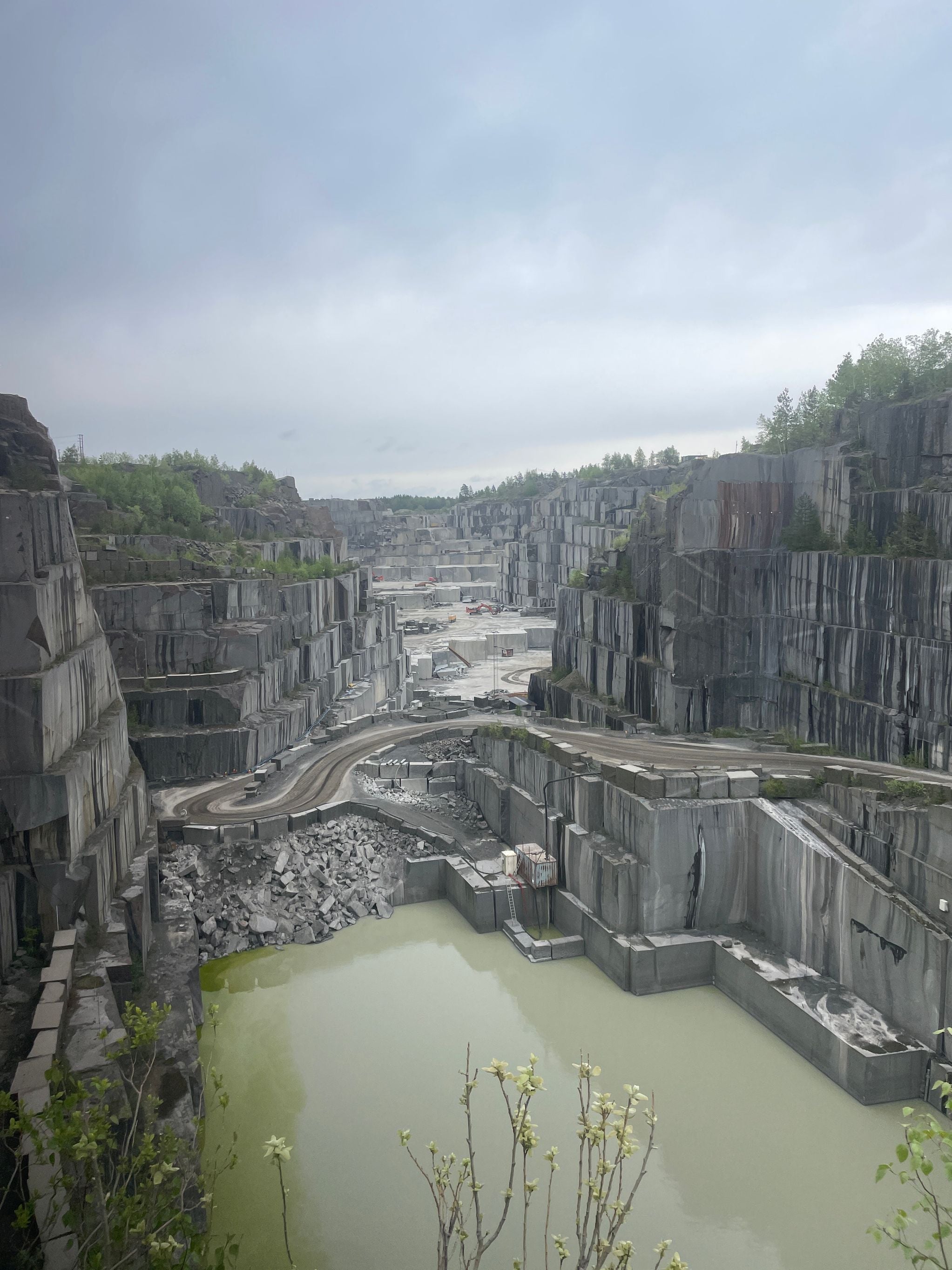 A large granite quarry with steep, terraced rock walls and a winding road leading through the site. Pools of greenish water collect at the bottom, and machinery is visible in the distance.