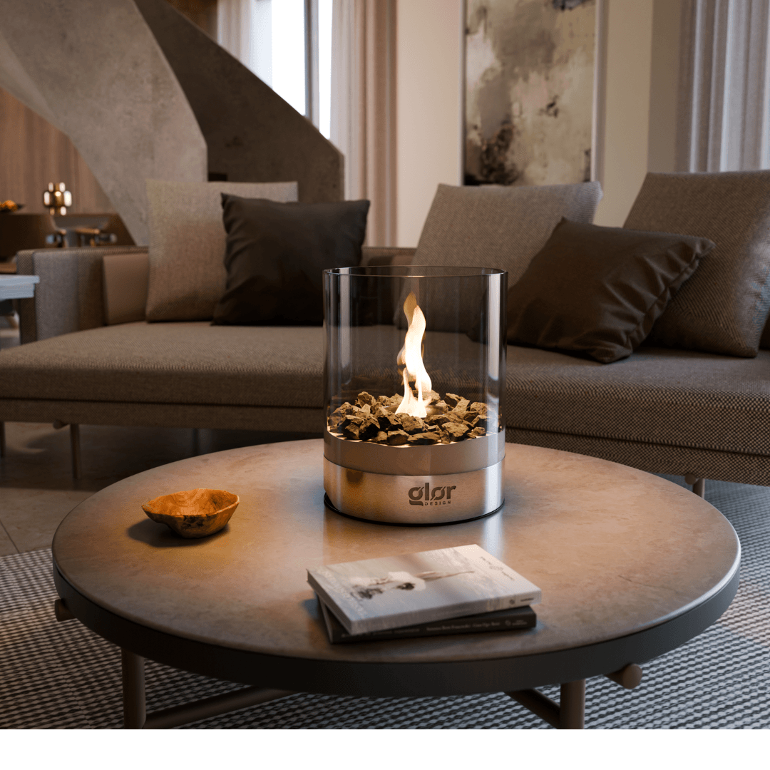 Modern tabletop fireplace with a cylindrical glass enclosure and decorative stones, placed on a round coffee table in a stylish living room with neutral-toned furniture and soft lighting.