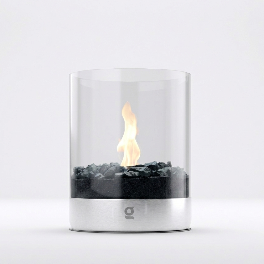 A modern tabletop fire pit with a clear glass cylinder, black decorative stones, and a visible flame, set against a clean white background.
